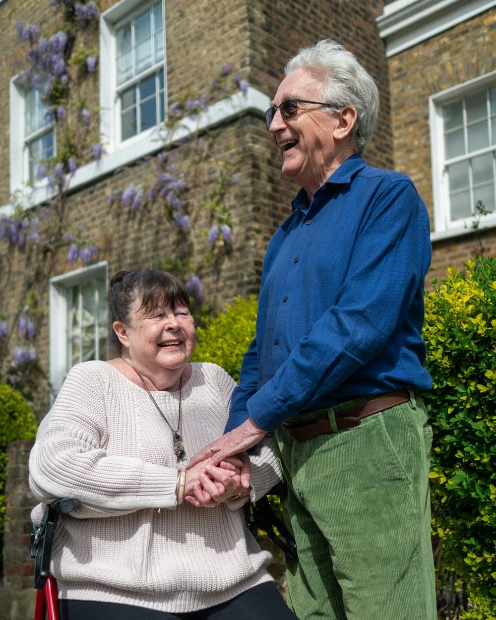 Two people laughing together outside a brick house, wisteria growing up the wall.