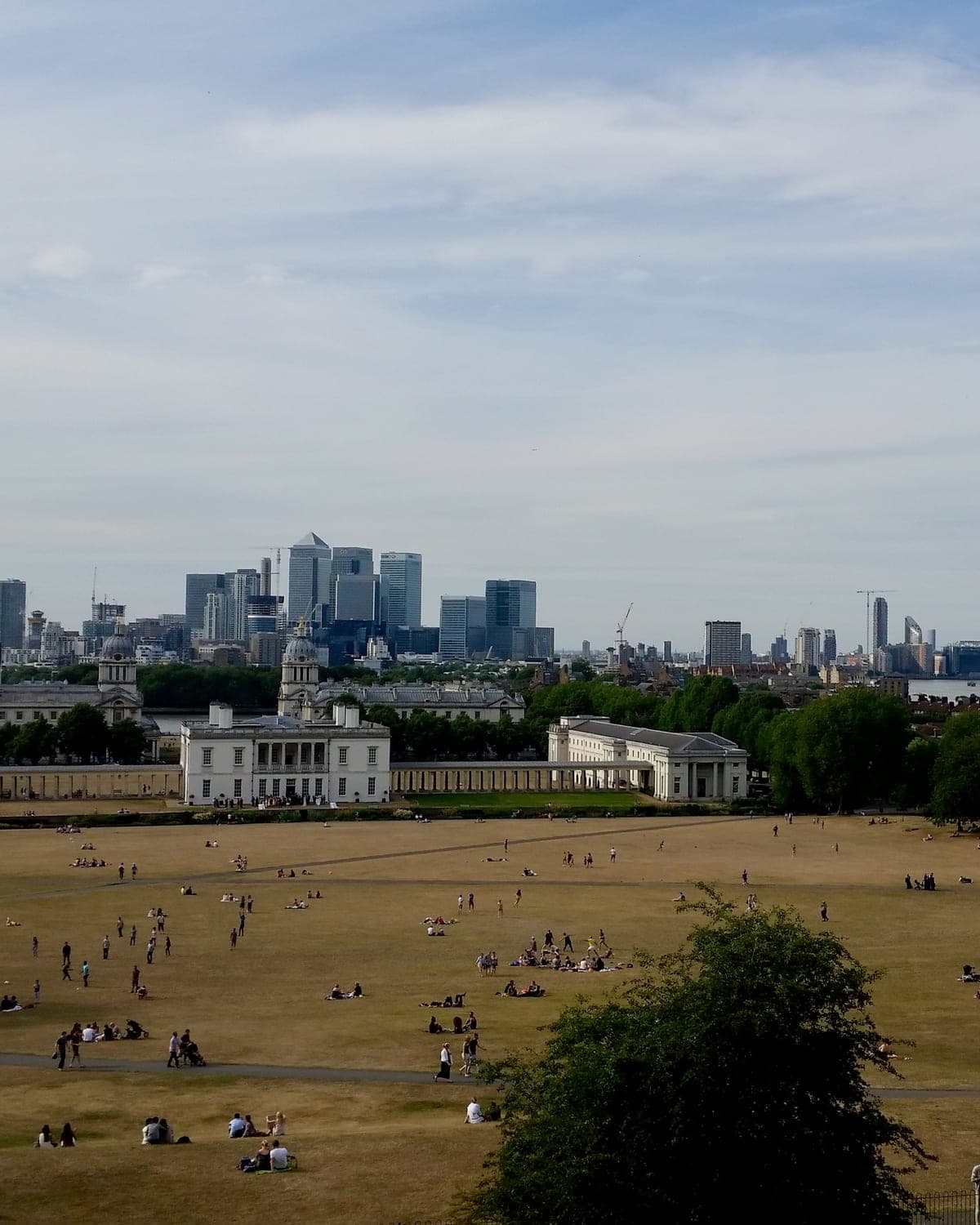 The view from the hill in Greenwich Park: people on the grass, the white Queen’s House below, and the towers of Canary Wharf across the river.