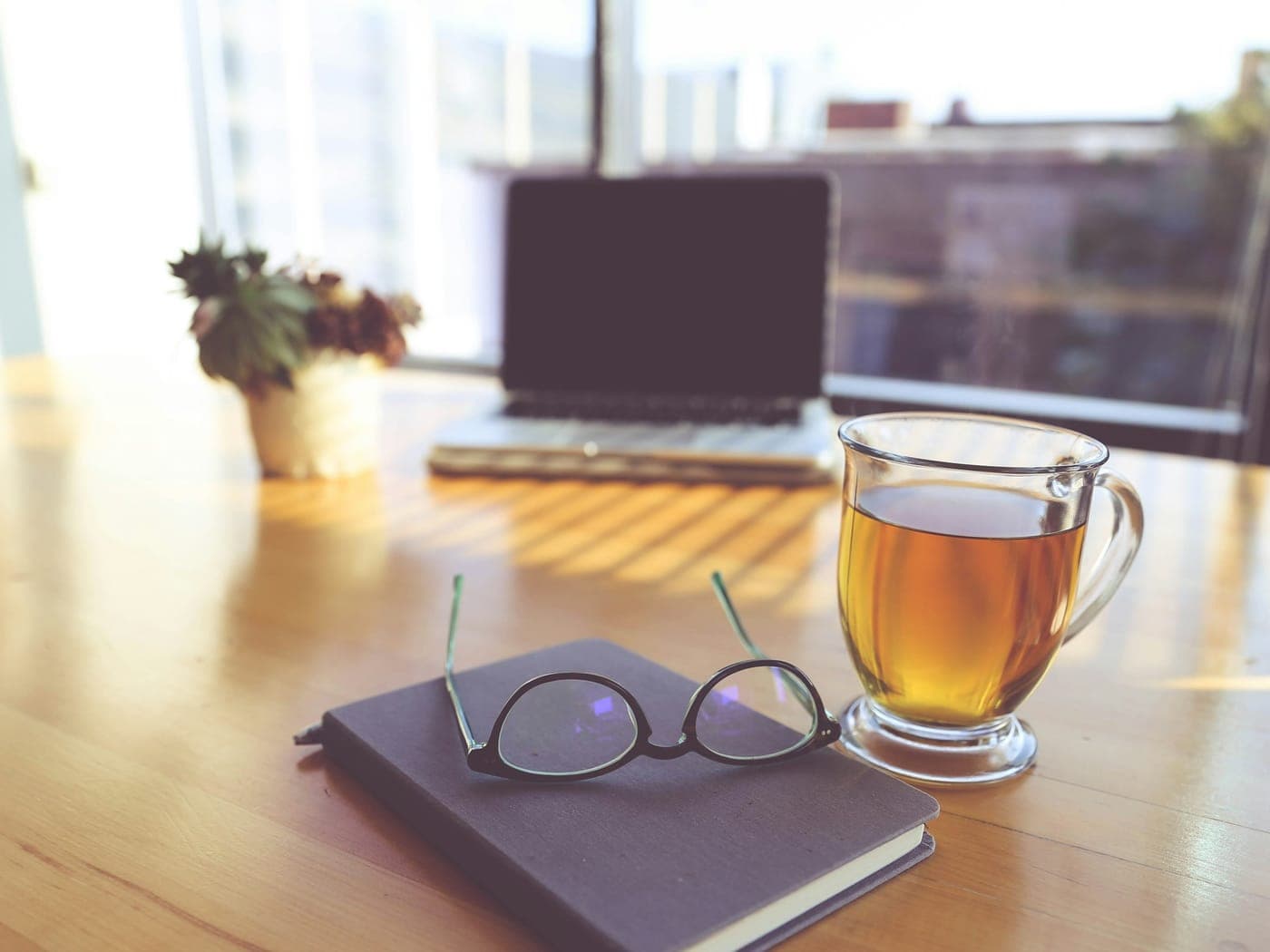 A notebook, a pair of reading glasses and a glass of tea on a table by a window.