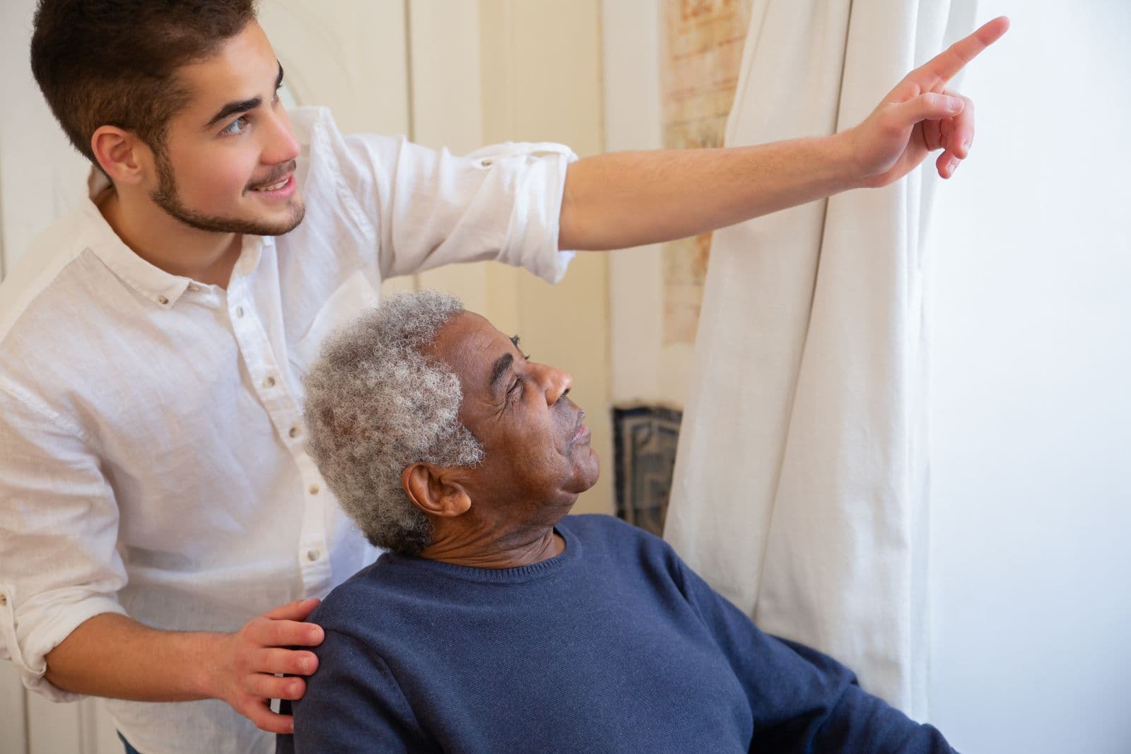 A carer rests a hand on an older man's shoulder and points out of the living room window, the two of them looking out together.